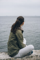 Side view of woman sitting on rock at beach against sky - CAVF55638