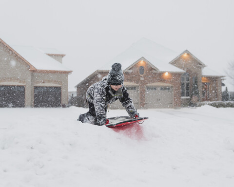 Junge rodelt bei Schneefall auf Schnee gegen Häuser, lizenzfreies Stockfoto