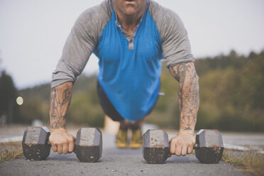 Low section of man with dumbbells doing push-ups on road at park - CAVF55675