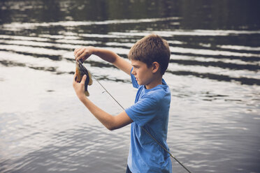 Side view of boy removing fish from fishing rod at lake - CAVF55764