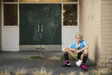 Portrait of girl in sports uniform holding water bottle while sitting on ball at yard - CAVF55766