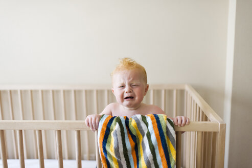 Baby boy crying while standing in crib against wall at home - CAVF55814