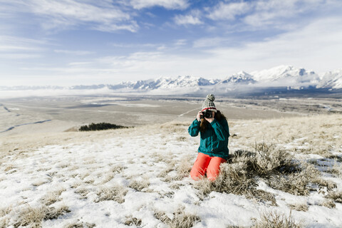 Wanderin fotografiert mit Kamera, während sie auf einem verschneiten Feld vor Bergen kniet, lizenzfreies Stockfoto