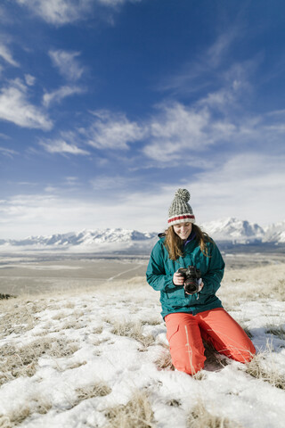 Female hiker looking at camera while kneeling on snowy field against mountains stock photo