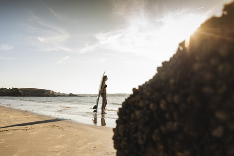 Frankreich, Bretagne, junge Frau mit Surfbrett am Meer stehend, lizenzfreies Stockfoto