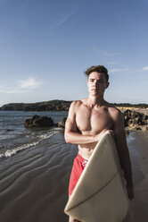 France, Brittany, young man carrying surfboard standing at the sea - UUF15899