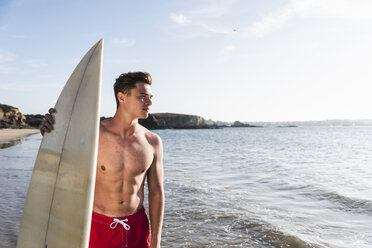 France, Brittany, young man carrying surfboard standing at the sea - UUF15901