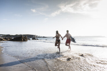 Frankreich, Bretagne, junges Paar mit Surfbrett läuft Hand in Hand im Meer - UUF15904