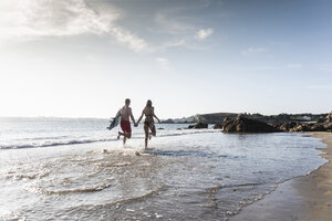 Frankreich, Bretagne, junges Paar mit Surfbrett läuft Hand in Hand im Meer - UUF15905
