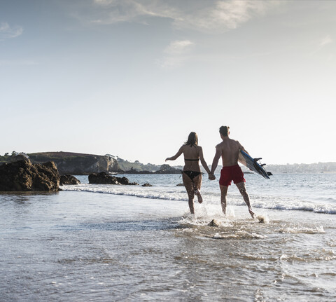 Frankreich, Bretagne, junges Paar mit Surfbrett läuft Hand in Hand im Meer, lizenzfreies Stockfoto