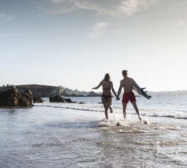 Frankreich, Bretagne, junges Paar mit Surfbrett läuft Hand in Hand im Meer - UUF15906