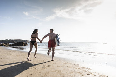 Frankreich, Bretagne, junges Paar mit Surfbrett läuft Hand in Hand im Meer - UUF15908