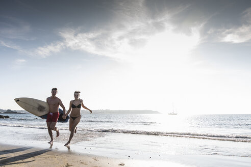 Frankreich, Bretagne, glückliches junges Paar mit Surfbrett läuft Hand in Hand im Meer - UUF15909