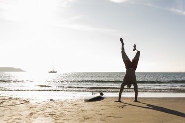 Frankreich, Bretagne, junger Mann macht einen Handstand am Strand neben einem Surfbrett - UUF15914