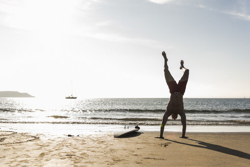 Frankreich, Bretagne, junger Mann macht einen Handstand am Strand neben einem Surfbrett - UUF15914