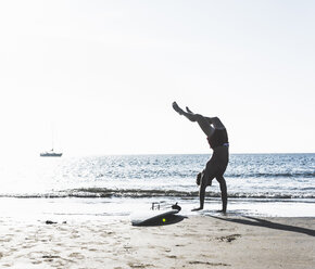 Frankreich, Bretagne, junger Mann macht einen Handstand am Strand neben einem Surfbrett - UUF15915