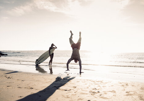 Frankreich, Bretagne, junger Mann macht einen Handstand und Frau hält Surfbrett am Strand - UUF15916