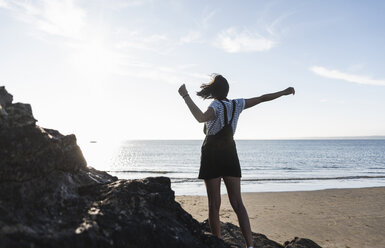 France, Brittany, rear view of young woman standing on rock at the beach at sunset - UUF15927