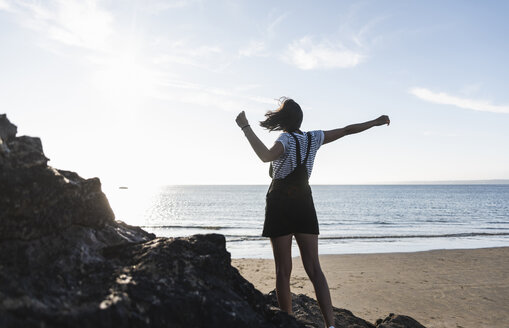 Frankreich, Bretagne, Rückansicht einer jungen Frau, die bei Sonnenuntergang auf einem Felsen am Strand steht - UUF15927