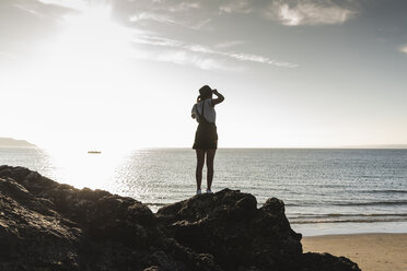 France, Brittany, rear view of young woman standing on rock at the beach at sunset - UUF15929