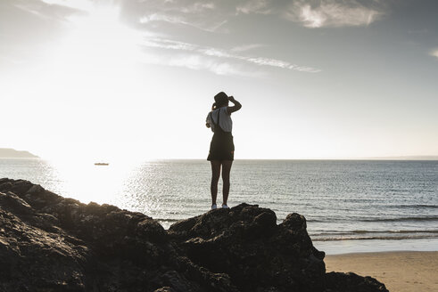 Frankreich, Bretagne, Rückansicht einer jungen Frau, die bei Sonnenuntergang auf einem Felsen am Strand steht - UUF15929