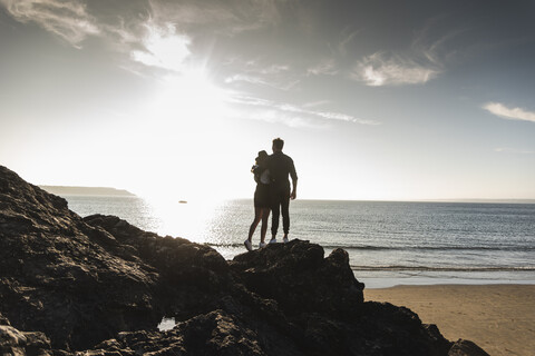 France, Brittany, rear view of young couple standing on rock at the beach at sunset stock photo