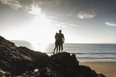 France, Brittany, rear view of young couple standing on rock at the beach at sunset - UUF15930