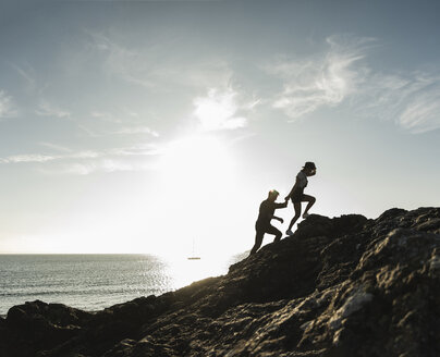 Frankreich, Bretagne, junges Paar klettert bei Sonnenuntergang auf einen Felsen am Strand - UUF15931