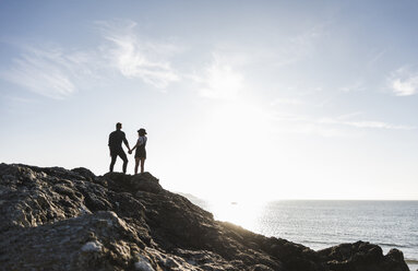 Frankreich, Bretagne, Rückansicht eines jungen Paares, das Hand in Hand auf einem Felsen am Strand bei Sonnenuntergang steht - UUF15932