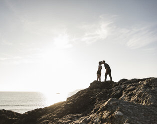 France, Brittany, young couple standing on rock at the beach at sunset kissing - UUF15933