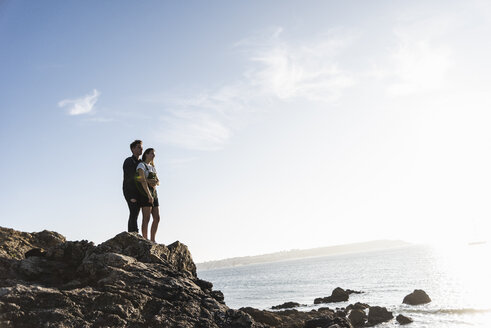 Frankreich, Bretagne, junges Paar steht bei Sonnenuntergang auf einem Felsen am Strand - UUF15934