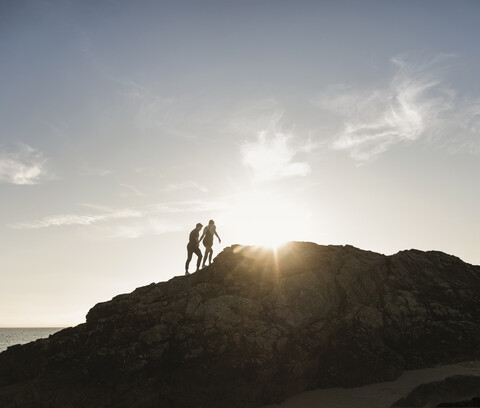 Frankreich, Bretagne, junges Paar klettert bei Sonnenuntergang auf einen Felsen am Strand, lizenzfreies Stockfoto