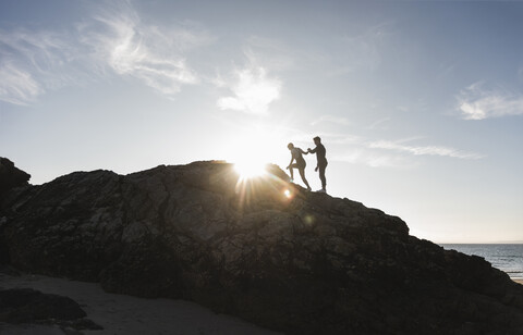 France, Brittany, young couple climbing on a rock at the beach at sunset stock photo