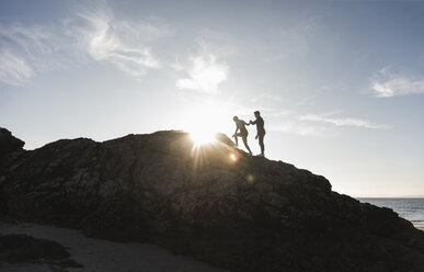 France, Brittany, young couple climbing on a rock at the beach at sunset - UUF15936