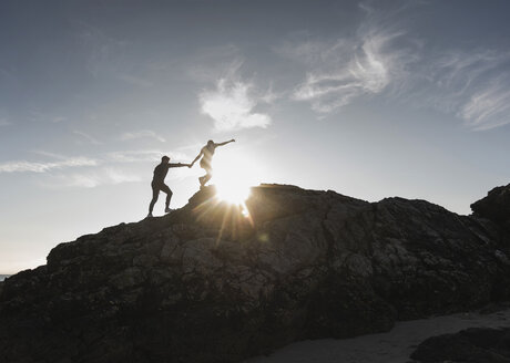 Frankreich, Bretagne, junges Paar klettert bei Sonnenuntergang auf einen Felsen - UUF15937