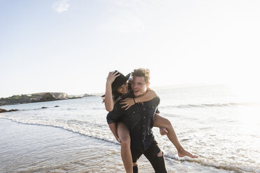 France, Brittany, happy young man carrying girlfriend piggyback at the beach - UUF15943