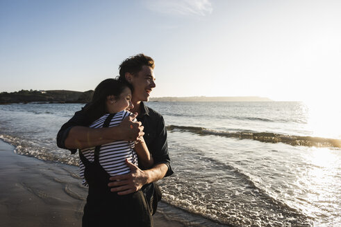 Frankreich, Bretagne, glückliches junges Paar, das sich bei Sonnenuntergang am Strand umarmt - UUF15948