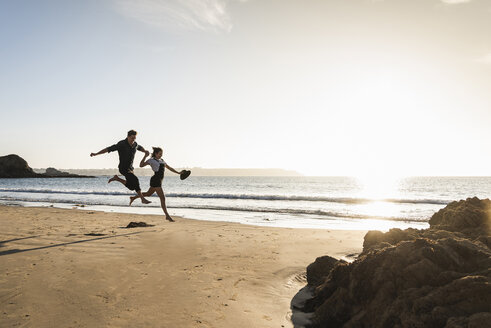 Frankreich, Bretagne, glückliches junges Paar springt bei Sonnenuntergang am Strand - UUF15950
