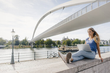 Netherlands, Maastricht, young woman sitting on a wall at the riverside with laptop - GUSF01401