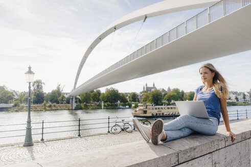 Niederlande, Maastricht, junge Frau sitzt mit Laptop auf einer Mauer am Flussufer - GUSF01401