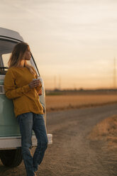 Young woman standing at camper van in rural landscape at sunset - GUSF01414