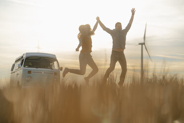 Exuberant couple at camper van in rural landscape with wind turbine in background - GUSF01418