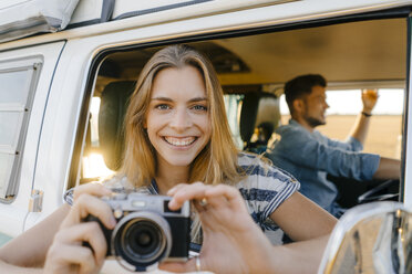 Portrait of happy woman with camera leaning out of window of a camper van with man driving - GUSF01424