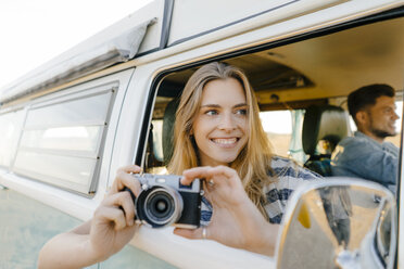 Hhappy woman with camera leaning out of window of a camper van with man driving - GUSF01425