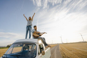 Happy couple with guitar on roof of a camper van in rural landscape - GUSF01442