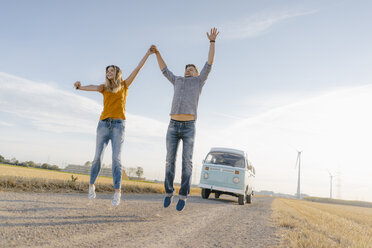 Exuberant couple jumping on dirt track at camper van in rural landscape - GUSF01453