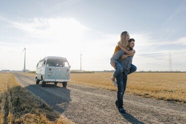 Young man carrying girlfriend piggyback on dirt track at camper van in rural landscape - GUSF01474
