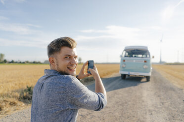 Lächelnder junger Mann, der mit seinem Handy ein Foto von einem Wohnmobil in einer ländlichen Landschaft macht - GUSF01476
