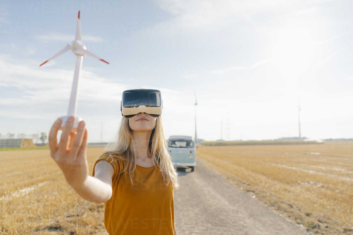 Junge Frau mit VR-Brille am Wohnmobil in ländlicher Landschaft hält Windrad-Modell,  lizenzfreies Stockfoto