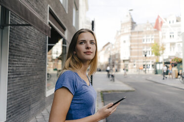 Niederlande, Maastricht, lächelnde junge Frau mit Mobiltelefon in der Stadt, die sich umschaut - GUSF01504